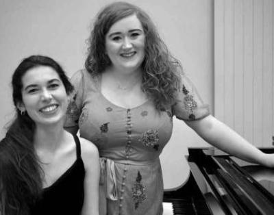 Black and white photo of Soprano Rosie Lavery and pianist Anna Michels stood next to a piano smiling at the camera.