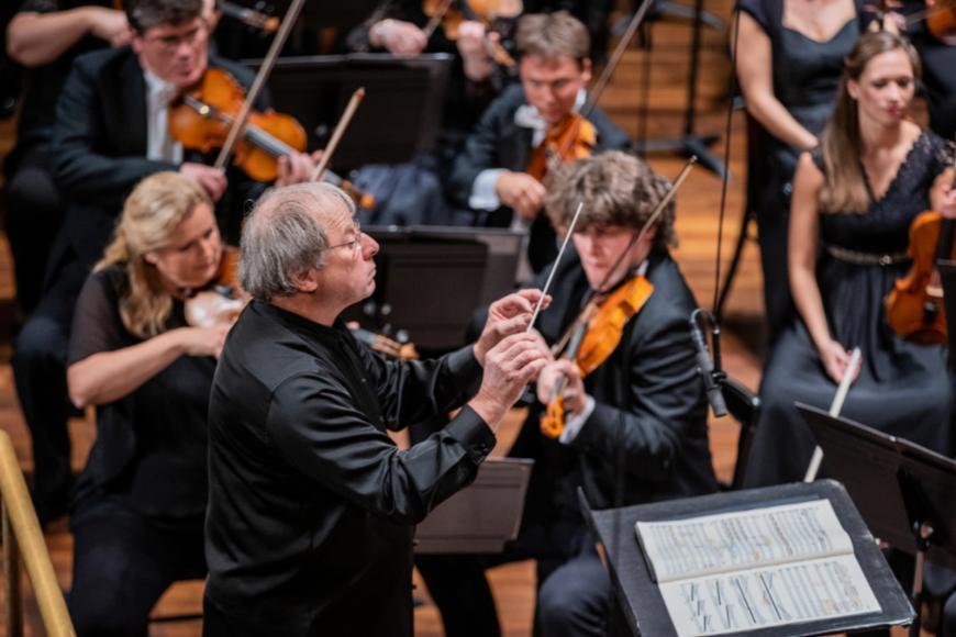 Conductor wears a black shirt and holds a baton. Sheet music is in front of him and he is surrounded by violinists performing.
