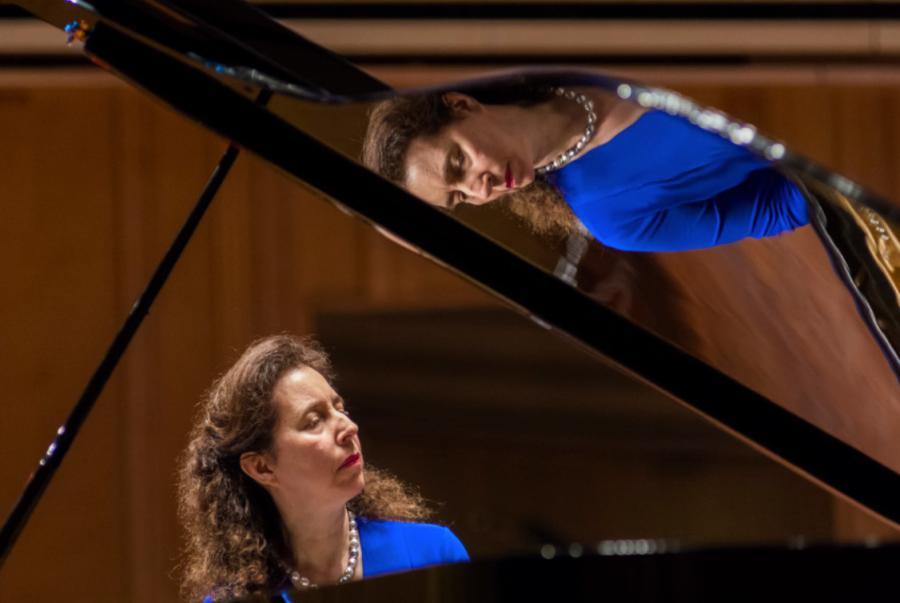 Pianist Angela Hewit wears a bright blue dress and sits at a grand piano with a raised lid. Her reflection is mirrored on the lid.