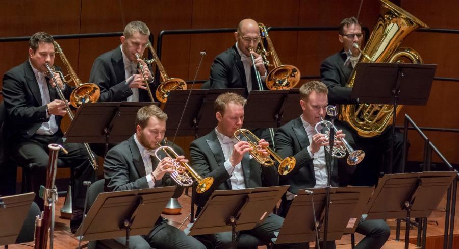 7 members of the orchestra are seated wearing formal attire and playing brass instruments while reading sheet music on stands.
